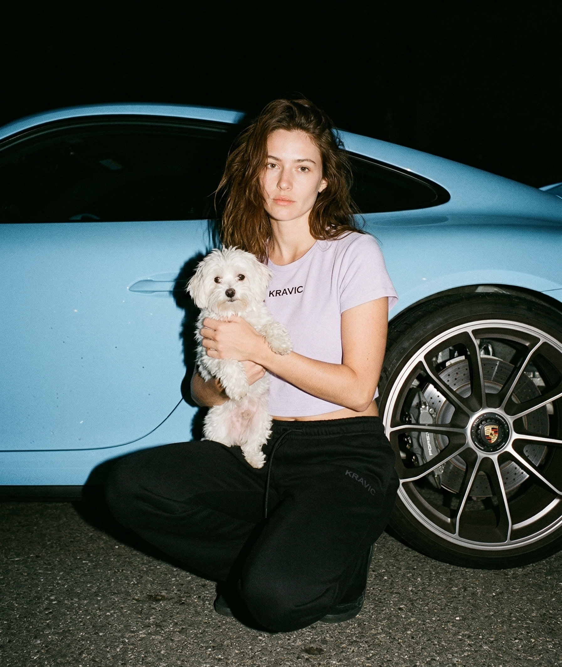 Woman holding a small white dog in front of a blue car at night.